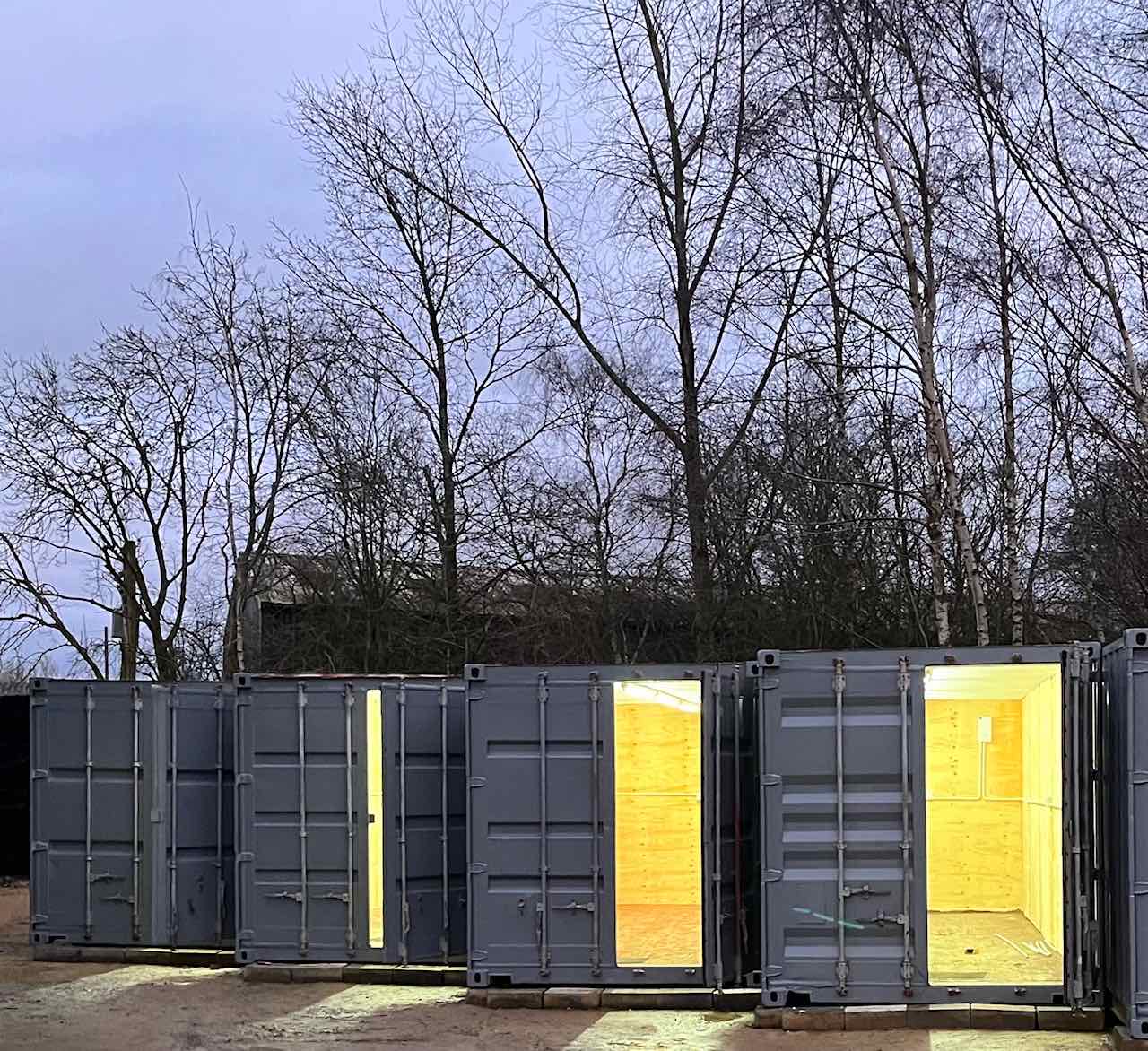 Row of grey GABA workshop containers at dusk, two with doors open showing lit, ply-lined interiors